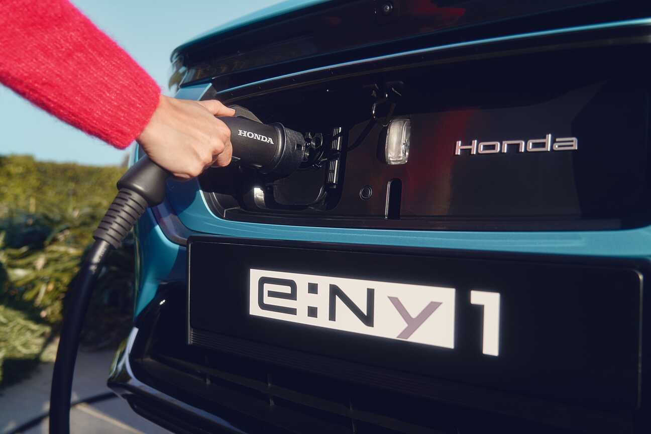 Close-up of a Honda electric vehicle being charged. The image shows a hand plugging in a charging cable to the rear of a blue Honda car. The license plate reads \"e:NY1\", indicating it's likely the Honda e:NY1 model.